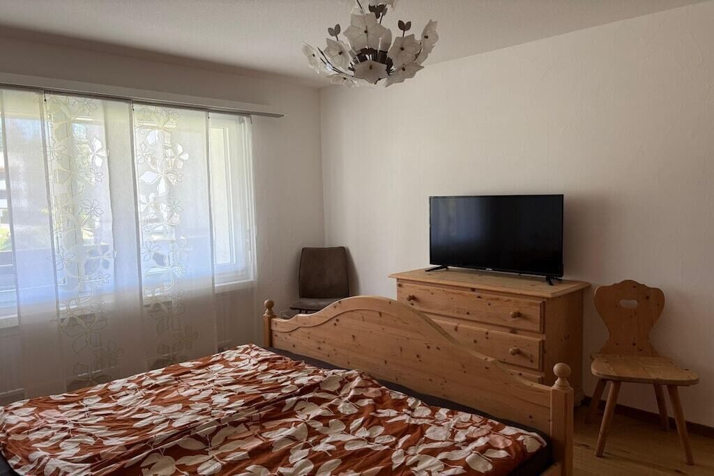 Ein Schlafzimmer mit einem Bett, einer Holzkommode, einem Fernseher, einem Sessel und einem Fenster mit Vorhängen.