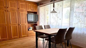 A dining area with a wooden table and chairs, a television on a shelf, and a window with floral curtains. - Holiday apartment in Laax with swimming pool (Laax)