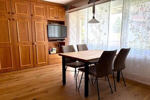 A dining area with a wooden table and chairs, a television on a shelf, and a window with floral curtains. - Holiday apartment in Laax with swimming pool (Laax)