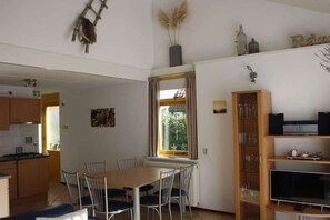 A dining area with a wooden table and chairs, a kitchen in the background, and a bookshelf with various items. - Holiday home with 3 bedrooms (Julianadorp aan Zee)