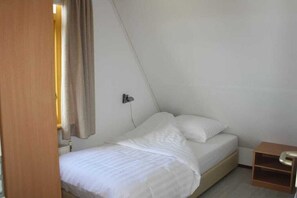 Habitació petita amb un llit individual, un panell de capçal de fusta, una tauleta de nit i una cortina en part oberta a l'esquerra.