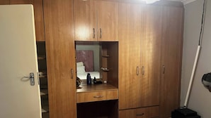 Un armario de madera con espejo, un escritorio con silla, y una mesita de noche con lámpara.