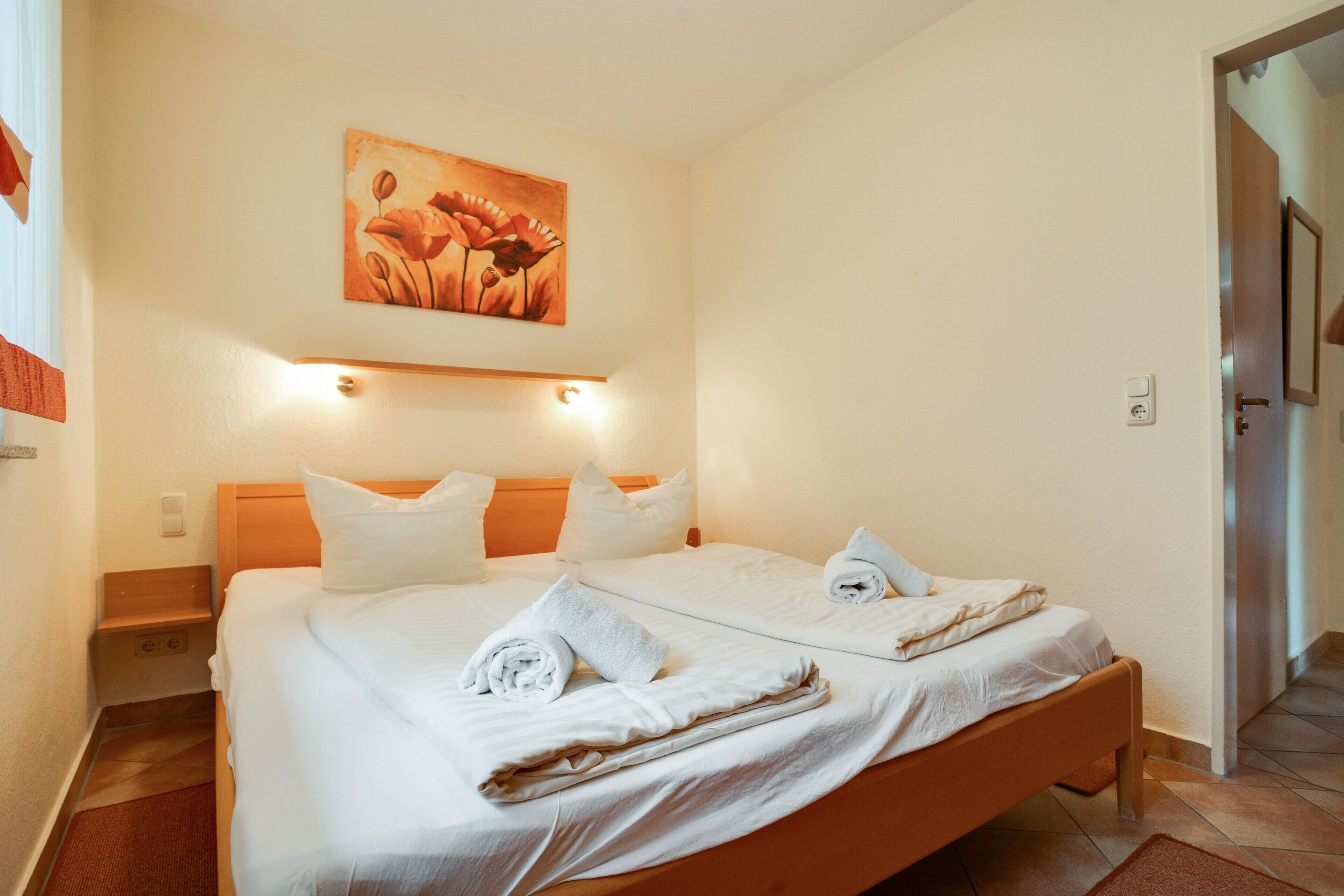 Ein Doppelbett mit weißen Leinen und Handtüchern, ein Holz-Kopfteil, ein Blumenbild und ein Lichtschalter an der Wand.
