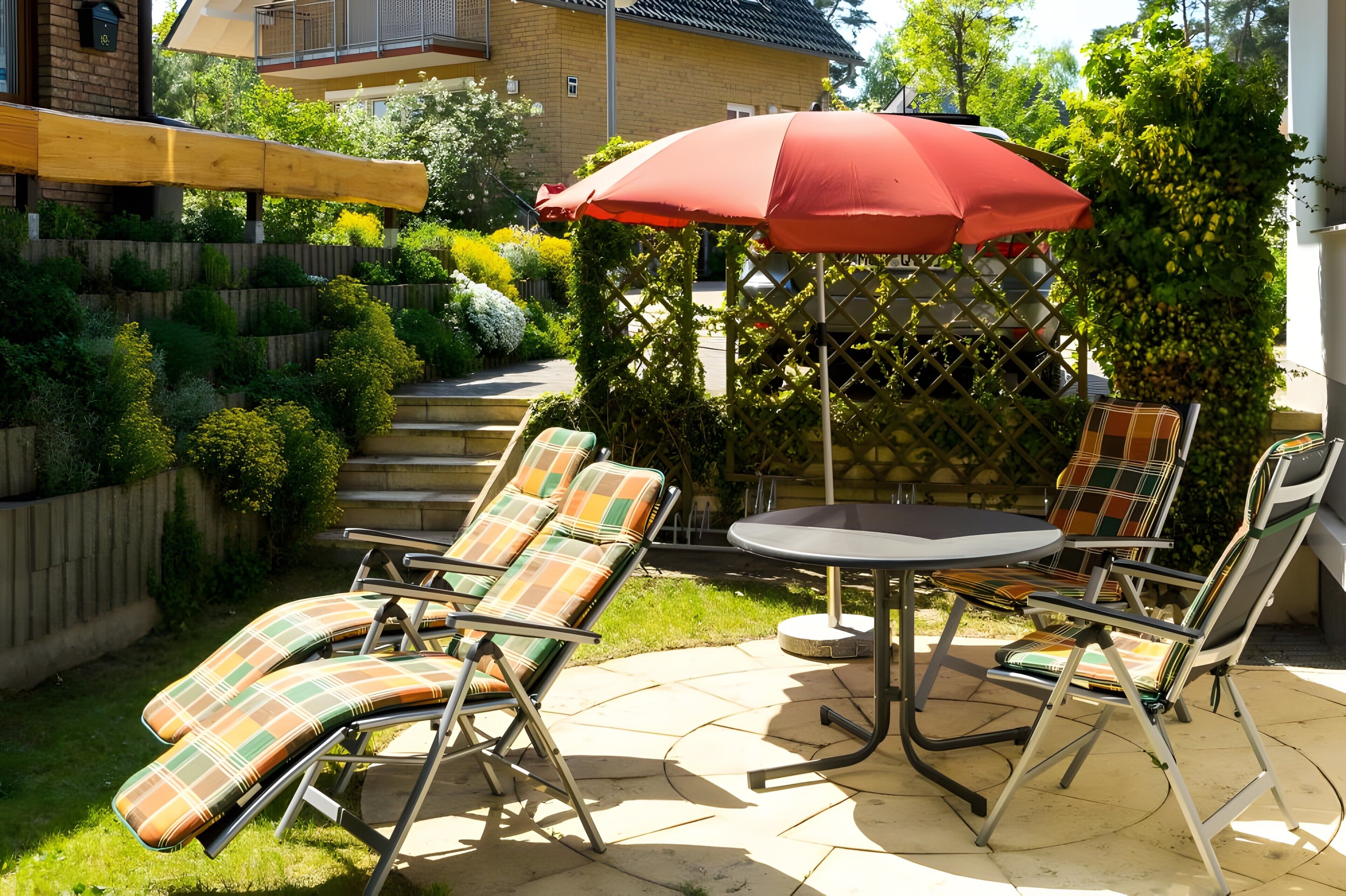 Eine Terrasse im Freien mit zwei Liegestühlen und einem Tisch, umgeben von einem Holenzaun und viel Grün.