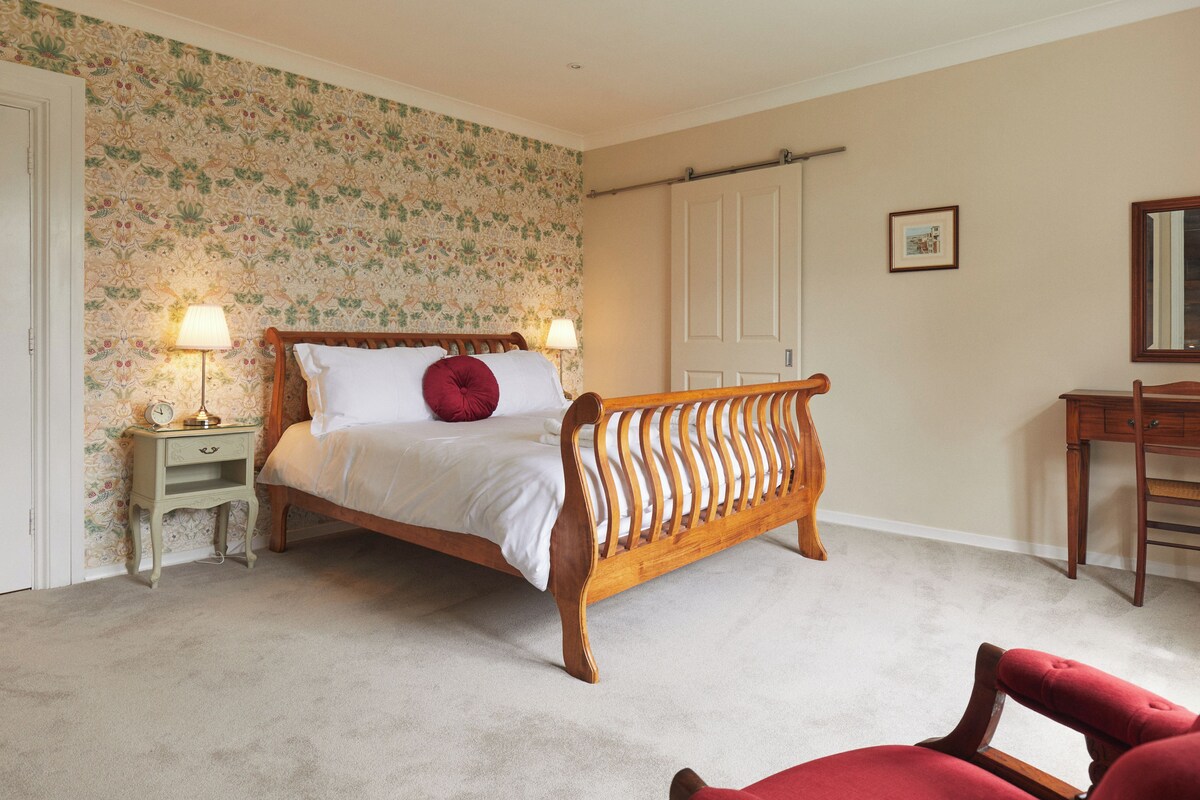 Un dormitori amb un llit de fusta, dues làmpades de nit, un coixí vermell, una cadira de fusta, un escriptori i un quadre amb marc a la paret.