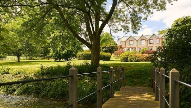 A wooden bridge leading to a building with a traditional architectural style, surrounded by lush greenery and a clear sky. - Lordington Park (Chichester)