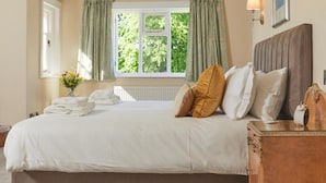 A neatly made bed with pillows, a bedside table with a lamp, and a window with curtains. - Lordington Park (Chichester)