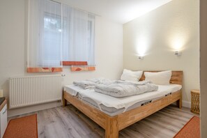 Ein Schlafzimmer mit einem hölzernen Bett, weißer Bettwäsche, einem Heizkörper, einem Fenster mit durchsichtigen Vorhängen und einer wandmontierten Lampe.
