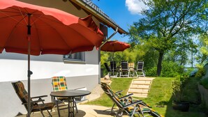 An outdoor patio with a table and chairs, a red umbrella, and a view of a garden with trees. - Müritzblick 3 - House Kranichruf - Lakefront Holiday Home with Sauna, Fireplace, Lake View & More (Röbel)