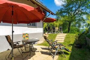 Eine Terrasse im Freien mit Tisch und Stühlen, einem roten Sonnenschirm und Blick auf einen Garten mit Bäumen.