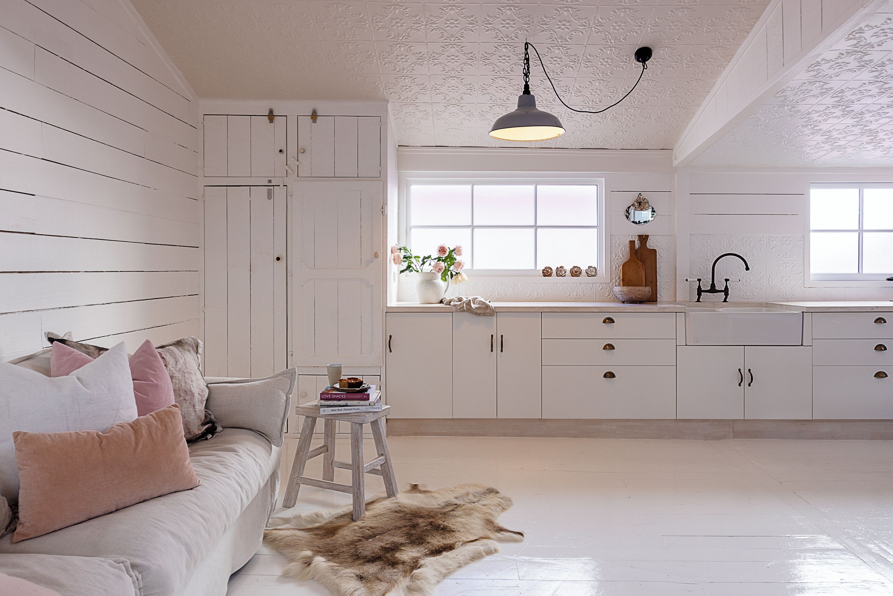 Ein Zimmer mit Holzboden in Weiß, einer weißen Couch mit Kissen, einem kleinen Hocker, einem Fellteppich, einem Fenster mit Ausblick und einer Küchenzeile mit Schränken und Spüle.