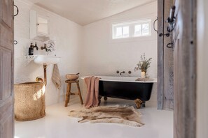 Een badkamer met een wit wastafel, een zwart bad, een houten krukje, een gevlochten mand en een vachtkleed.