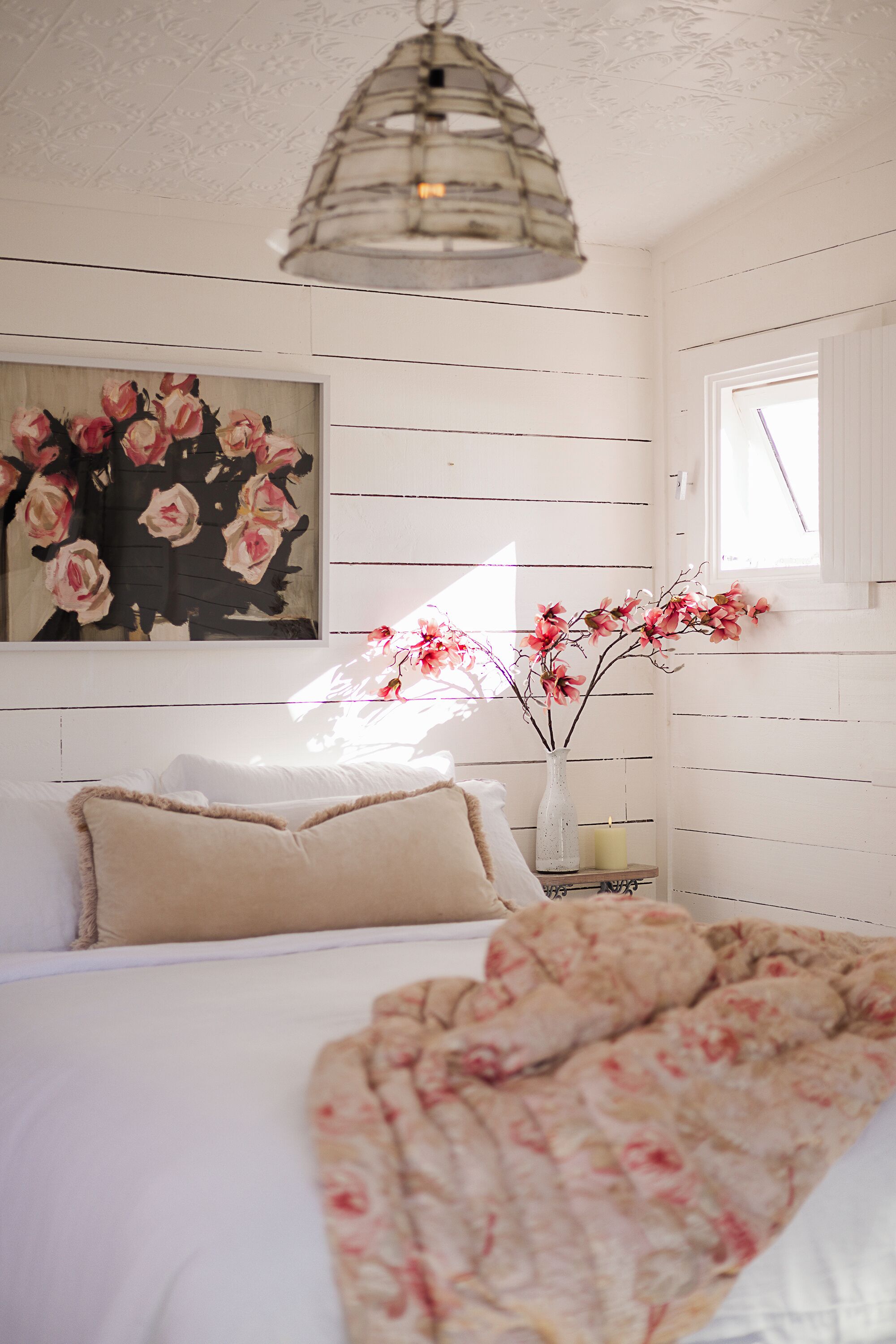 A bedroom with a bed, pillows, a vase with flowers, a painting, and a window.