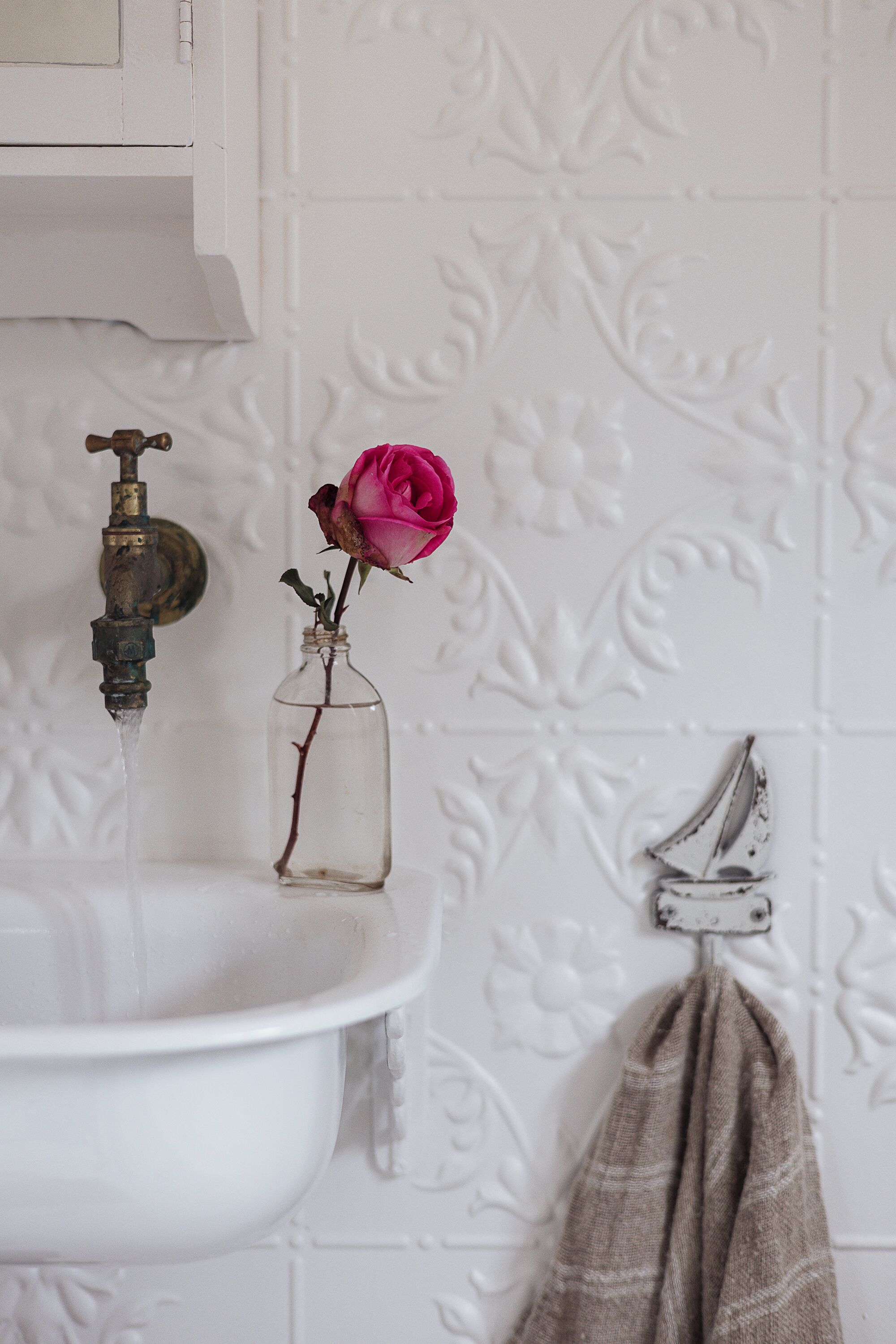 A bathroom with a white sink, a rose in a glass jar, and a towel hanging on a rack.