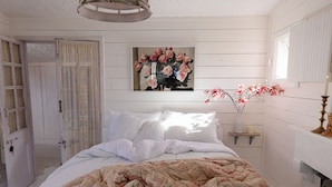 A bedroom with a bed, a painting, a vase with flowers, and a door.