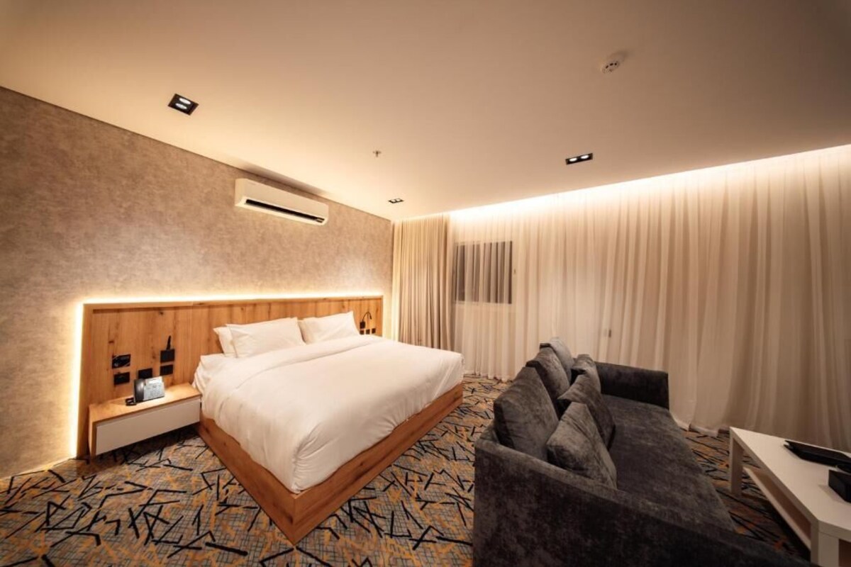 Quarto de hotel com uma cama grande, um sofá, uma mesa de cabeceira e ar condicionado fixado na parede.