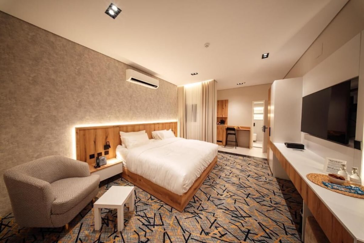 Quarto de hotel com cama, cadeira, escrivaninha, televisão e ar condicionado.
