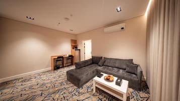 Una habitación de hotel moderna con un sofá seccional gris, una pequeña mesa blanca con un cuenco de frutas, un escritorio con sillas y una alfombra estampada.
