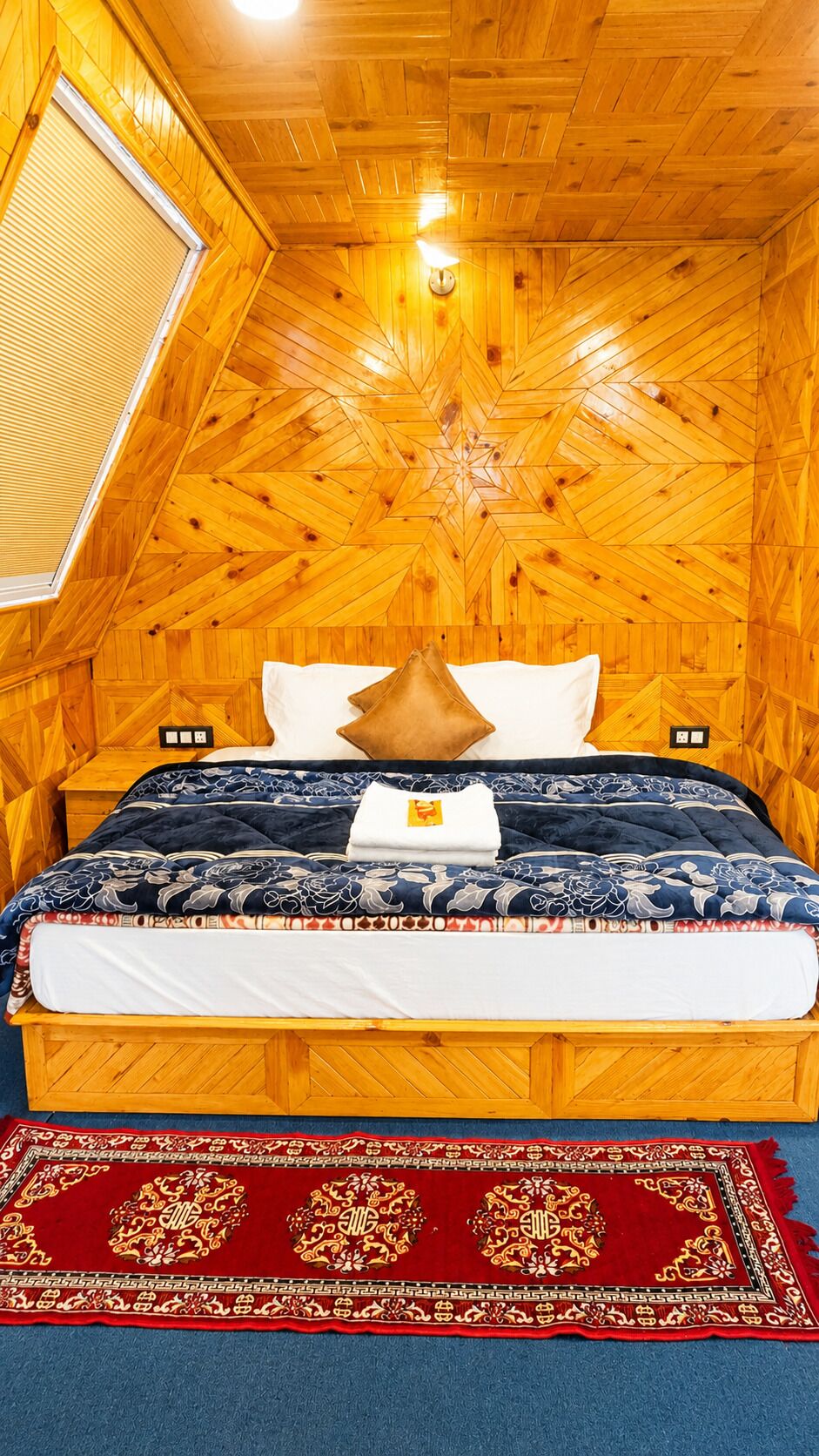 Una habitación con paneles de madera, una cama, un ventanal y un tapete rojo con estampado.