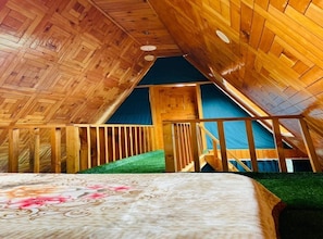 An attic bedroom with a wooden sloped ceiling, a bed with a floral bedspread, and a green artificial turf floor. - Latos Chapter Sajwar (Banjar)
