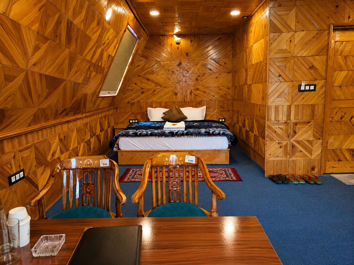 Ein Zimmer mit Holzwänden, einem Bett, zwei Stühlen, einem Tisch und einem Paar Hausschuhen auf dem Boden.