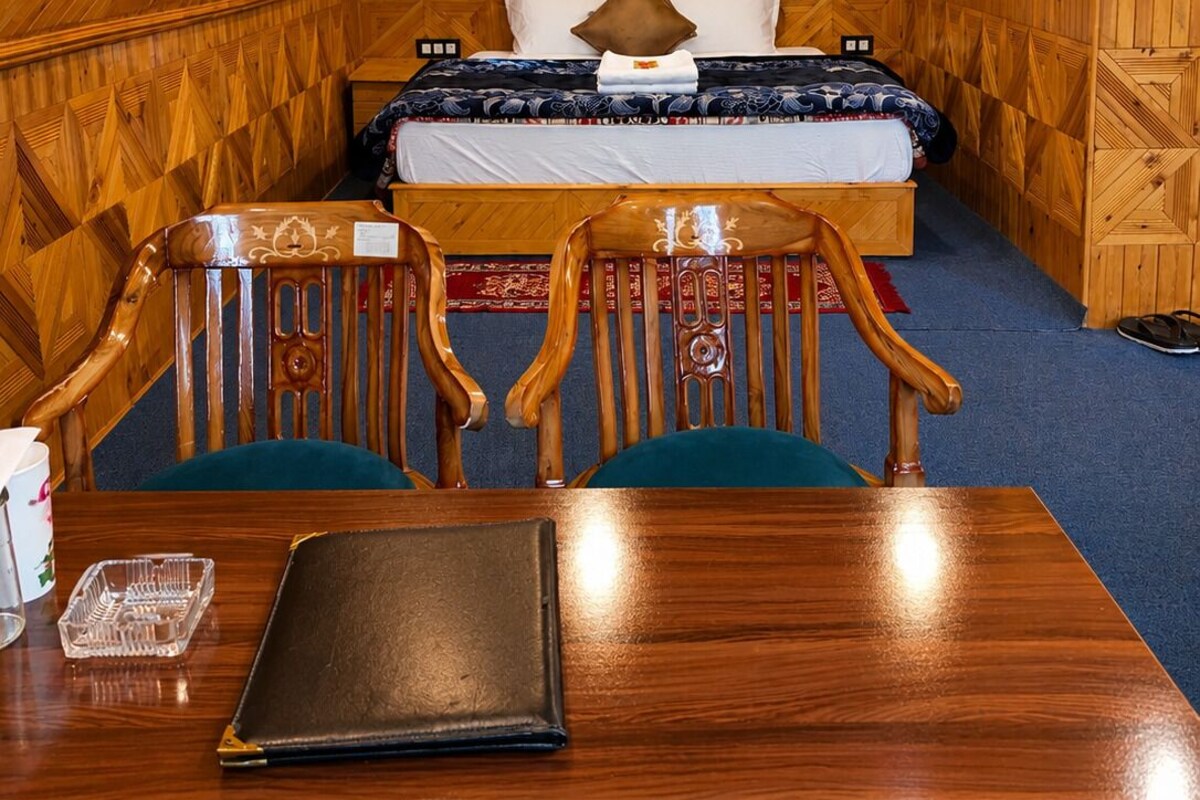 A wooden-paneled room with a bed, two chairs, a table, and a small table lamp.