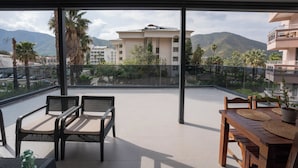 Varanda com mesa e cadeiras, um vaso de planta e vista para um prédio e montanhas.