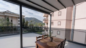 Uma varanda com mesa e cadeiras de madeira, um vaso com planta e vista para montanhas e edifícios.
