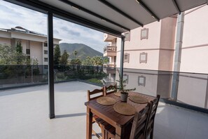 Een balkon met een houten tafel en stoelen, een plant in een pot en uitzicht op een berg en gebouwen.