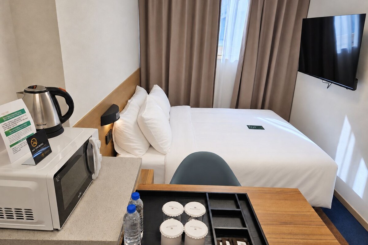 Een hotelkamer met een bed, een televisie, een waterkoker, een magnetron, en een dienblad met waterflessen en verpakkingen.