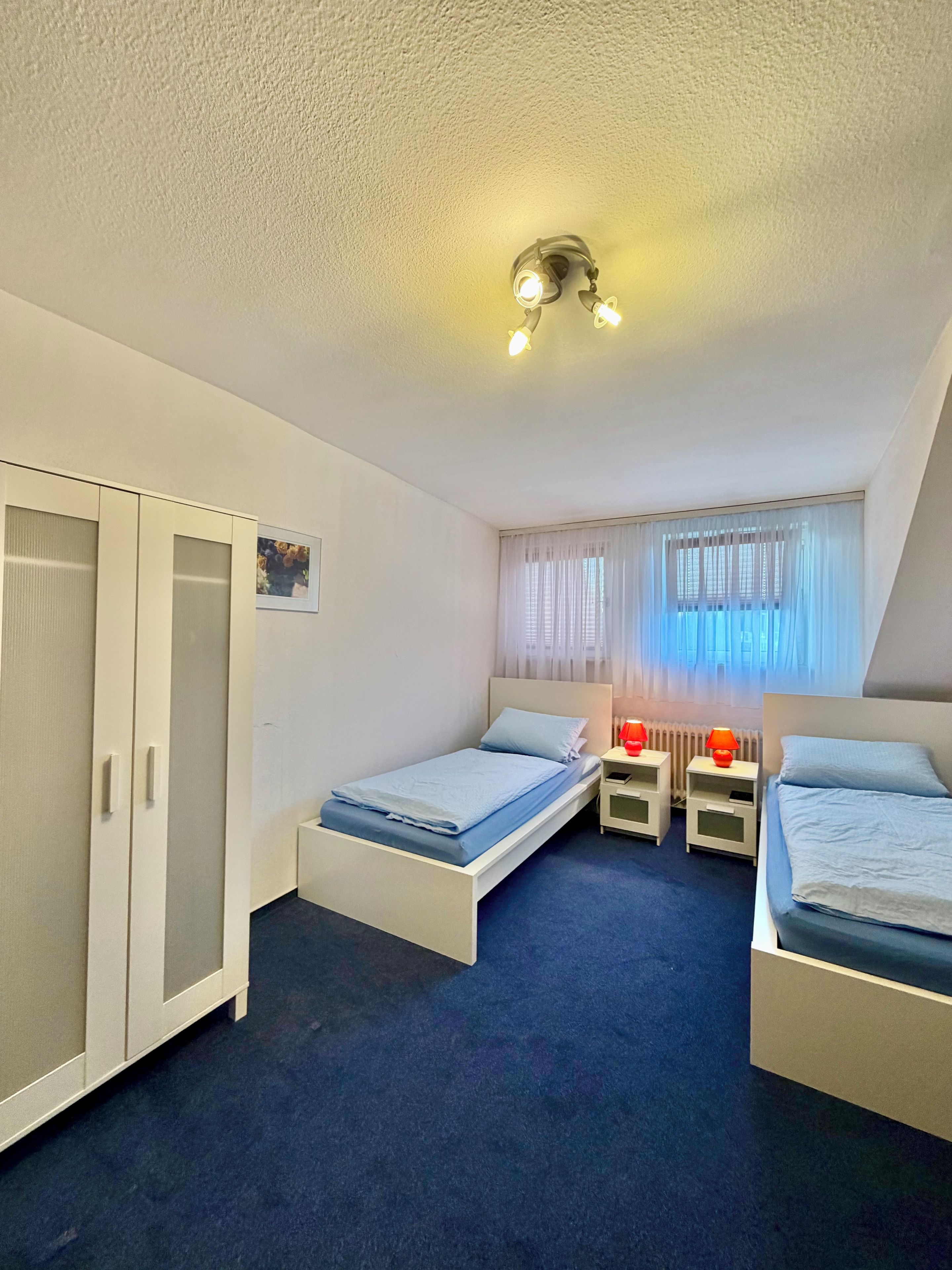 A room with two single beds, a wardrobe, a window with curtains, and a ceiling light fixture.