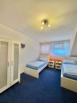 Una habitación con dos camas individuales, un armario, una ventana con cortinas y una lámpara en el techo.