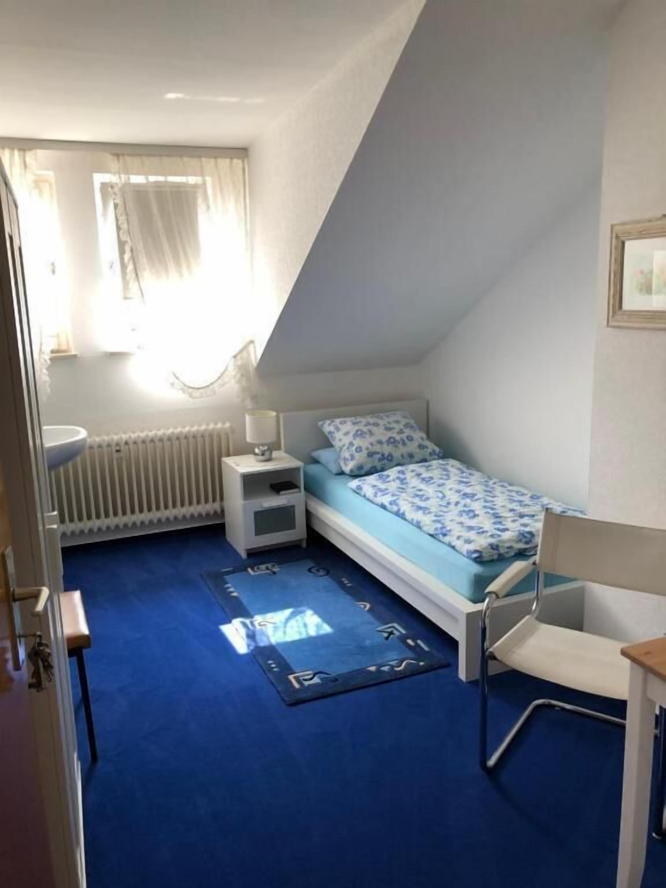 A small, single-bed room with a sloped ceiling, a bedside table, a chair, and a radiator.