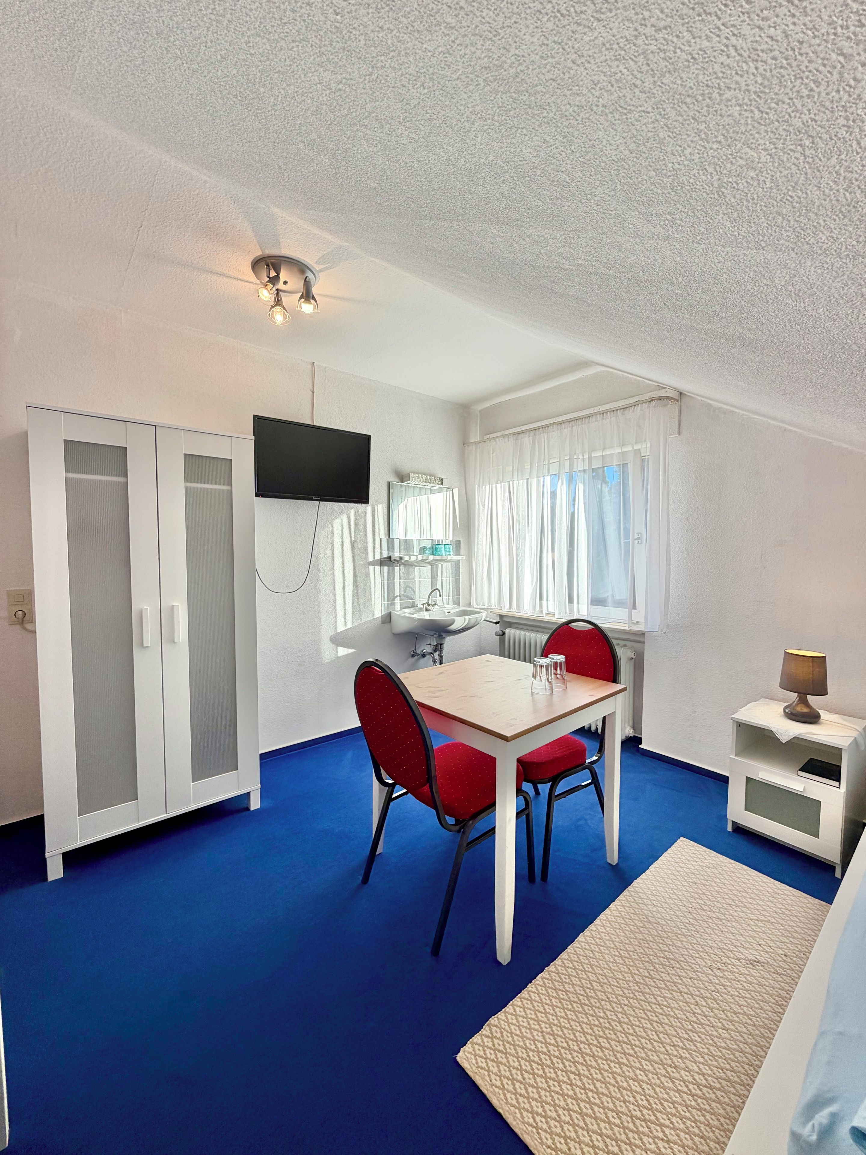 A room with a dining table, chairs, a TV mounted on the wall, a window with curtains, and a white wardrobe.