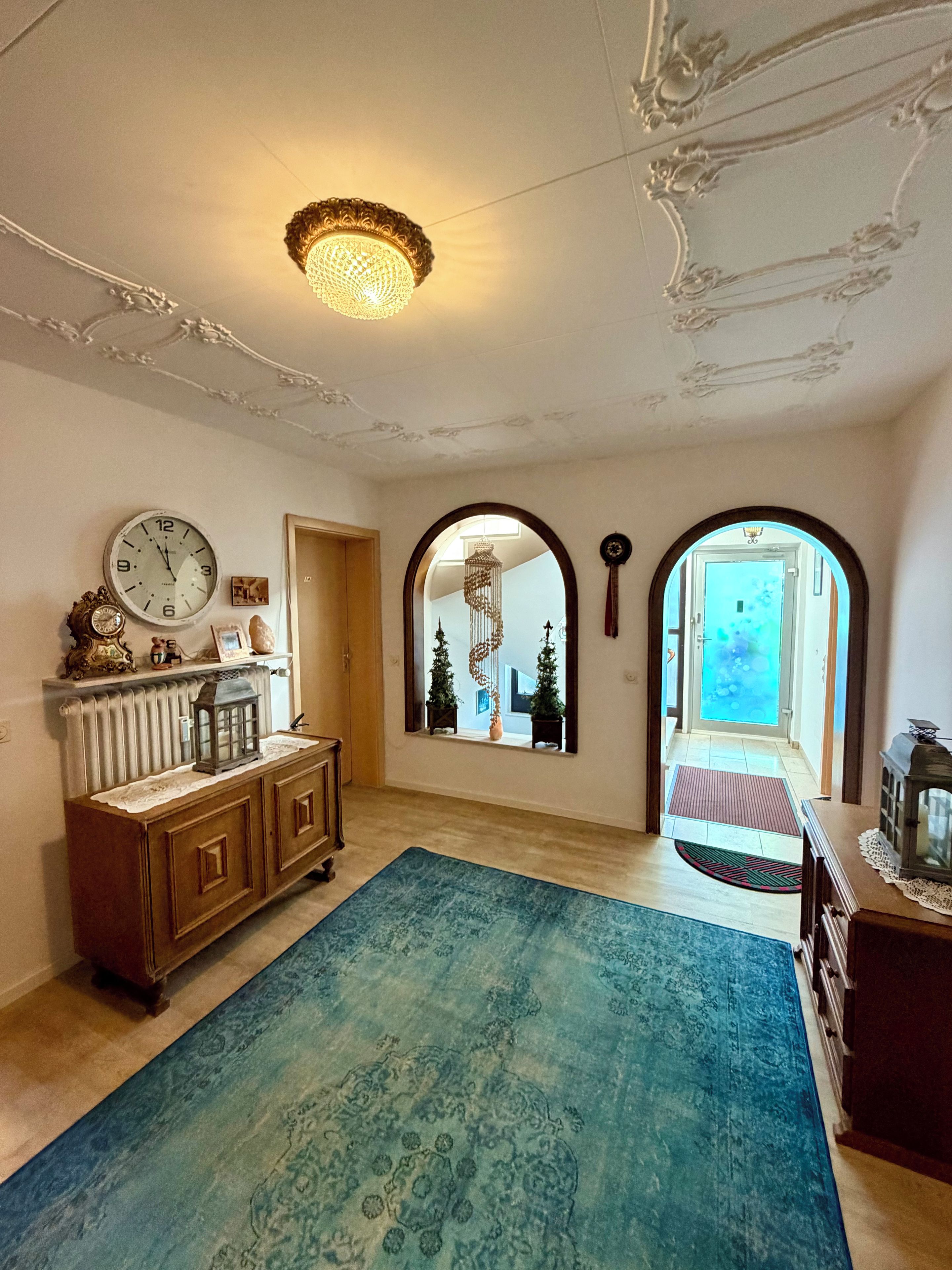 Une pièce avec un grand tapis bleu, un meuble en bois orné d’une horloge et un lustre.