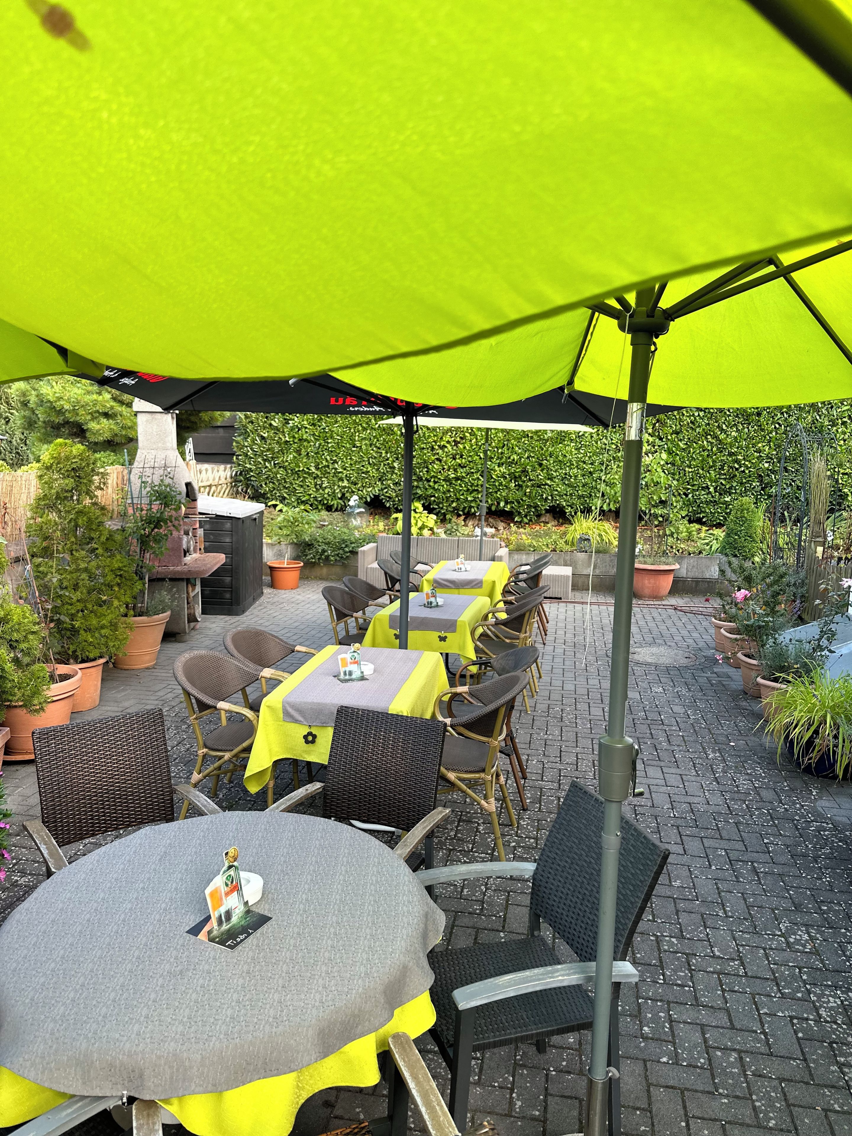 An outdoor patio with tables and chairs, some covered by umbrellas, and potted plants around.