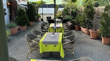 An outdoor patio with tables and chairs, covered by umbrellas, and surrounded by potted plants.