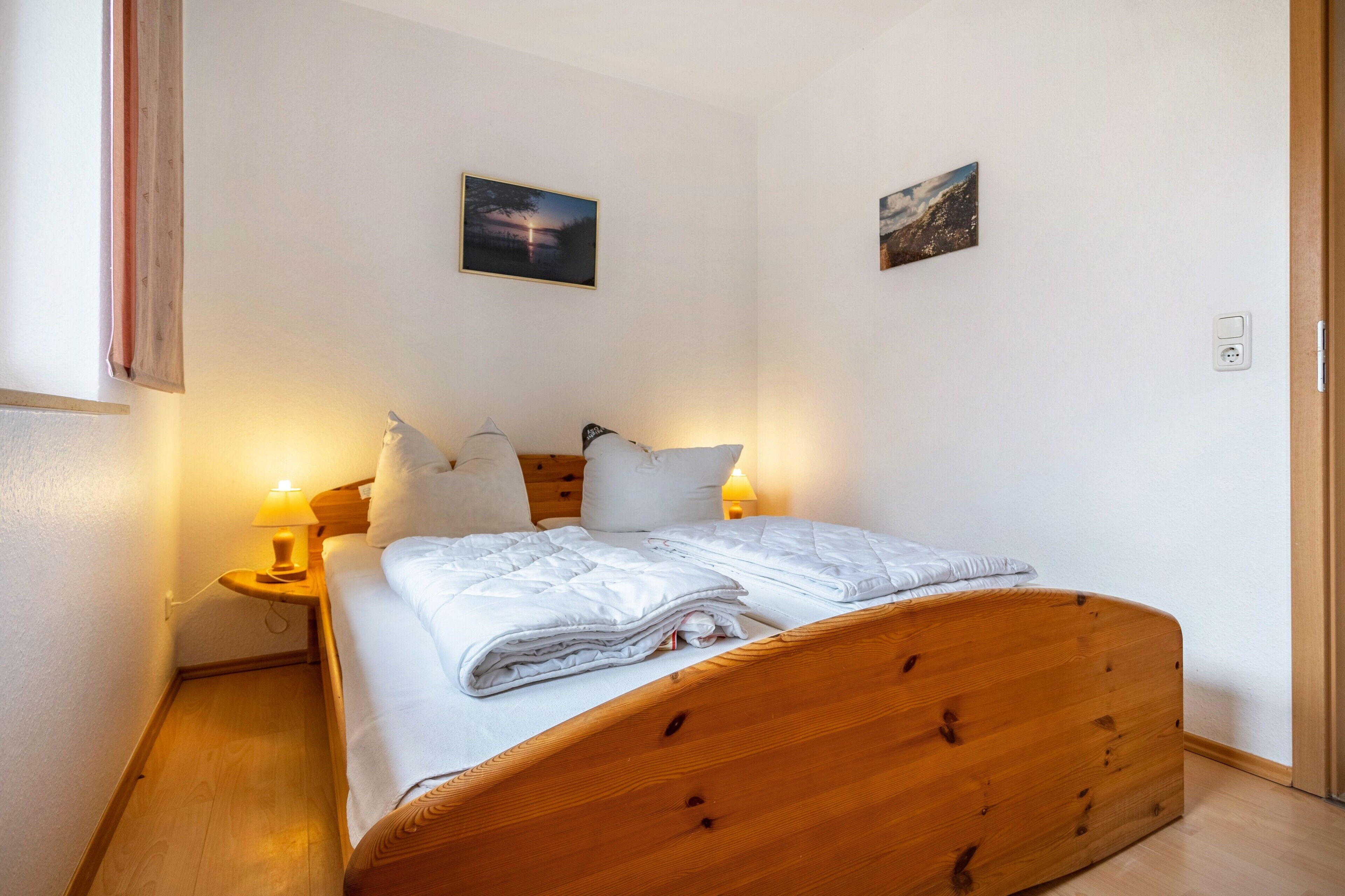 Een eenpersoonsbed met houten hoofdbord in een kamer met witte muren, twee nachtkastjes met lampen en twee ingelijste foto's aan de muur.