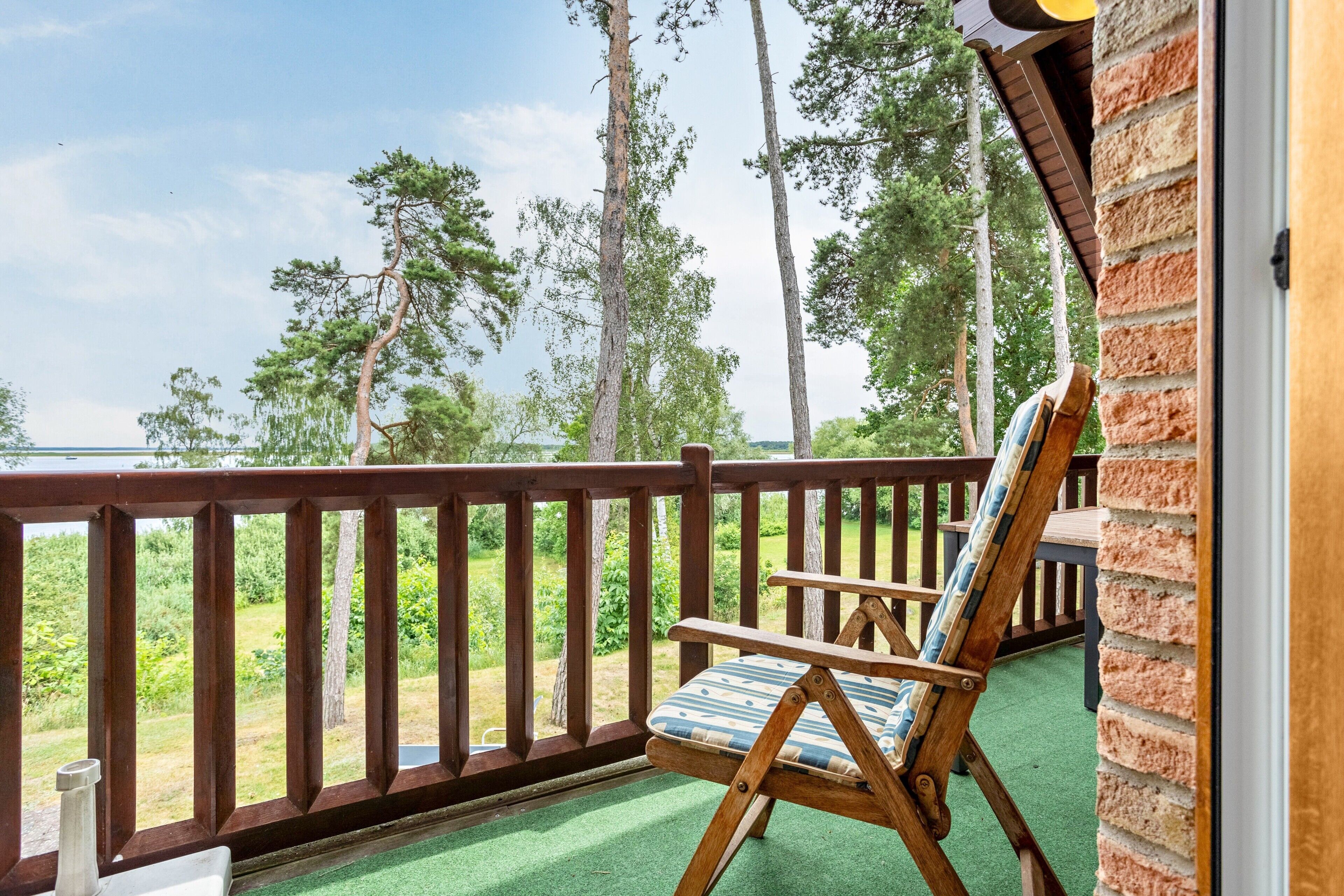 Eine Holzterrasse mit einem einzelnen Schaukelstuhl, mit Blick auf eine Wiese mit Bäumen und einem Gewässer.