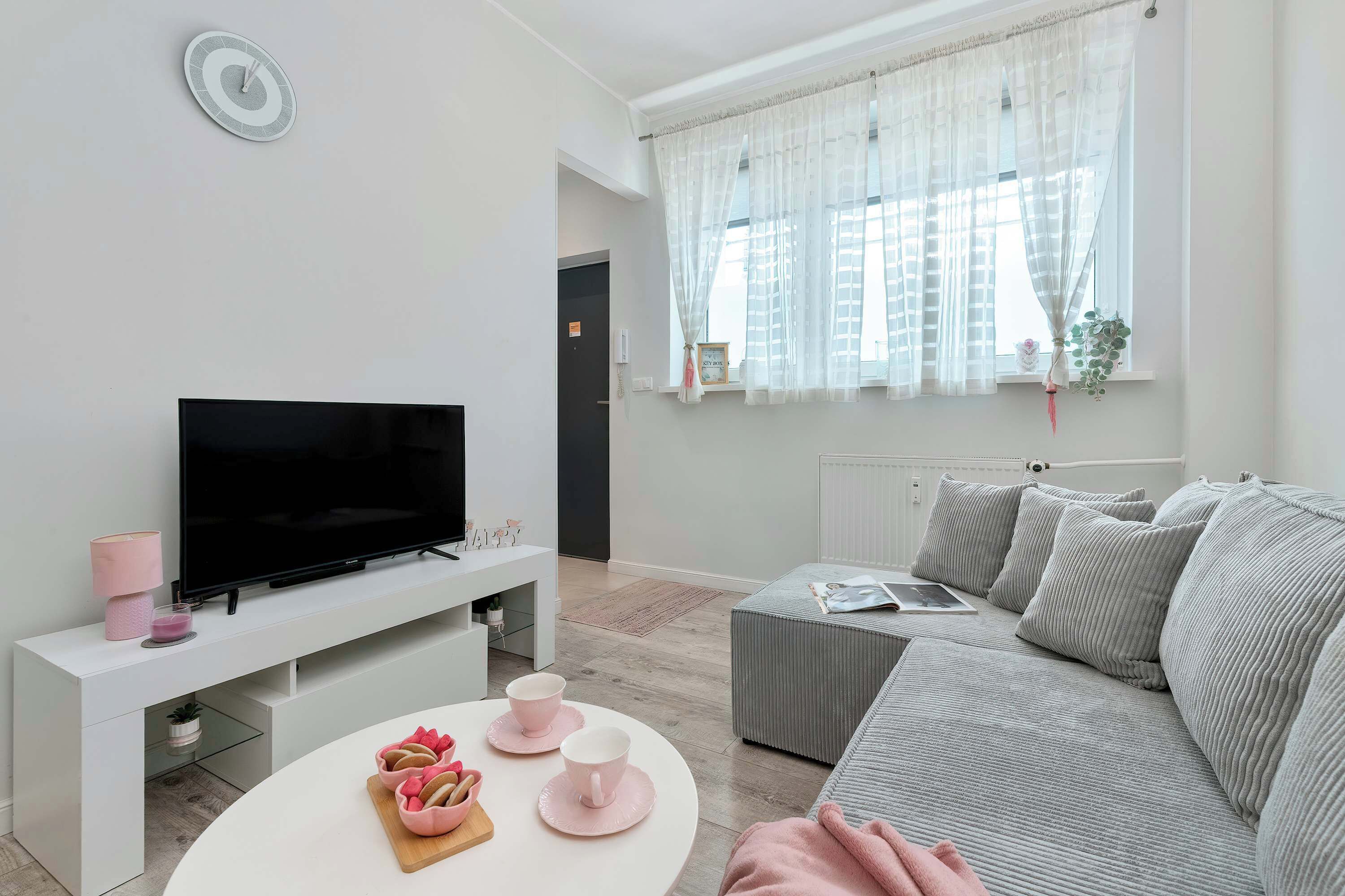 A modern living room with a flat-screen TV on a white stand, a grey sofa, and a white coffee table with pink cups and a wooden board.