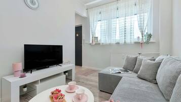 A modern living room with a flat-screen TV on a white stand, a grey sofa, and a white coffee table with pink cups and a wooden board.