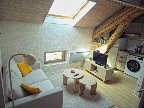 Ett kompakt bostadsutrymme med ett sovloft, en soffa med gula kuddar, ett runt soffbord, en tv, en tvättmaskin och ett takfönster.