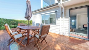 Une terrasse en bois avec une table ronde et des chaises, un parasol, et une vue sur l’intérieur de la maison à travers une porte vitrée.