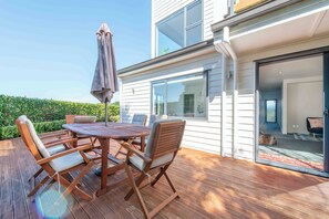 Une terrasse en bois avec une table ronde et des chaises, un parasol, et une vue sur l’intérieur de la maison à travers une porte vitrée.