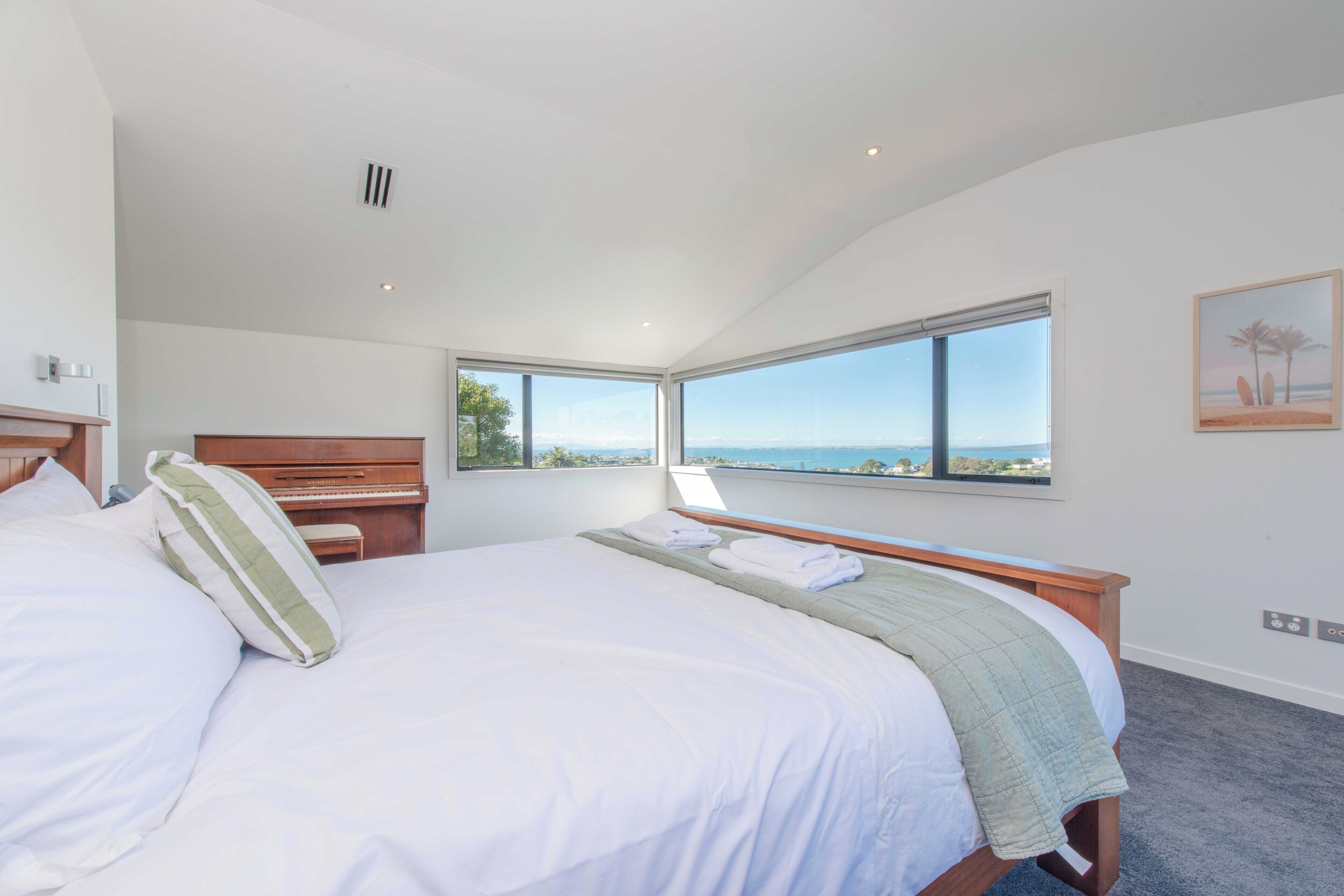 A bedroom with a large bed, a piano, a view of the sea, and a painting on the wall.