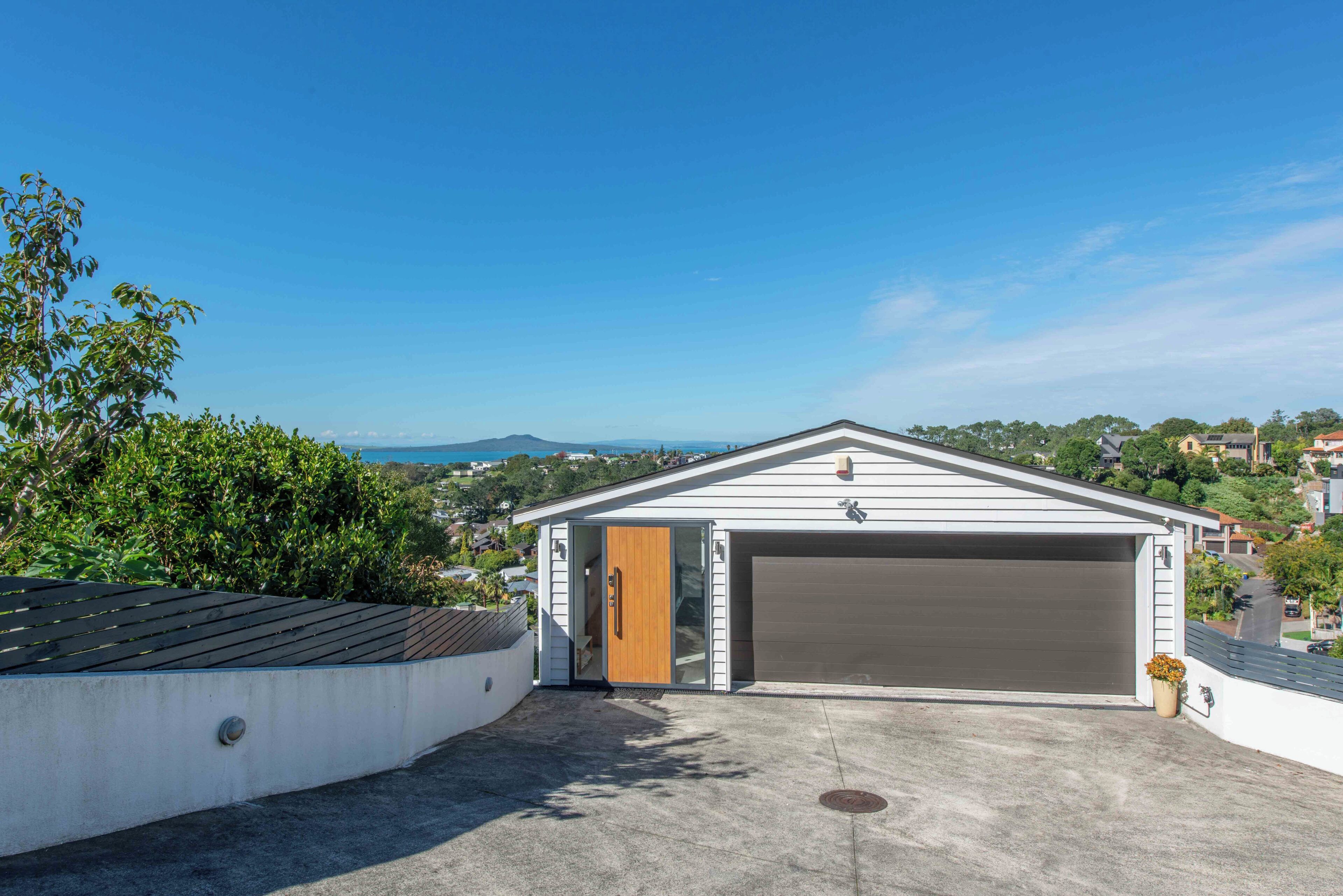Un garage moderno con porta in legno, vialetto in cemento e vista aperta su una zona residenziale e colline sullo sfondo.