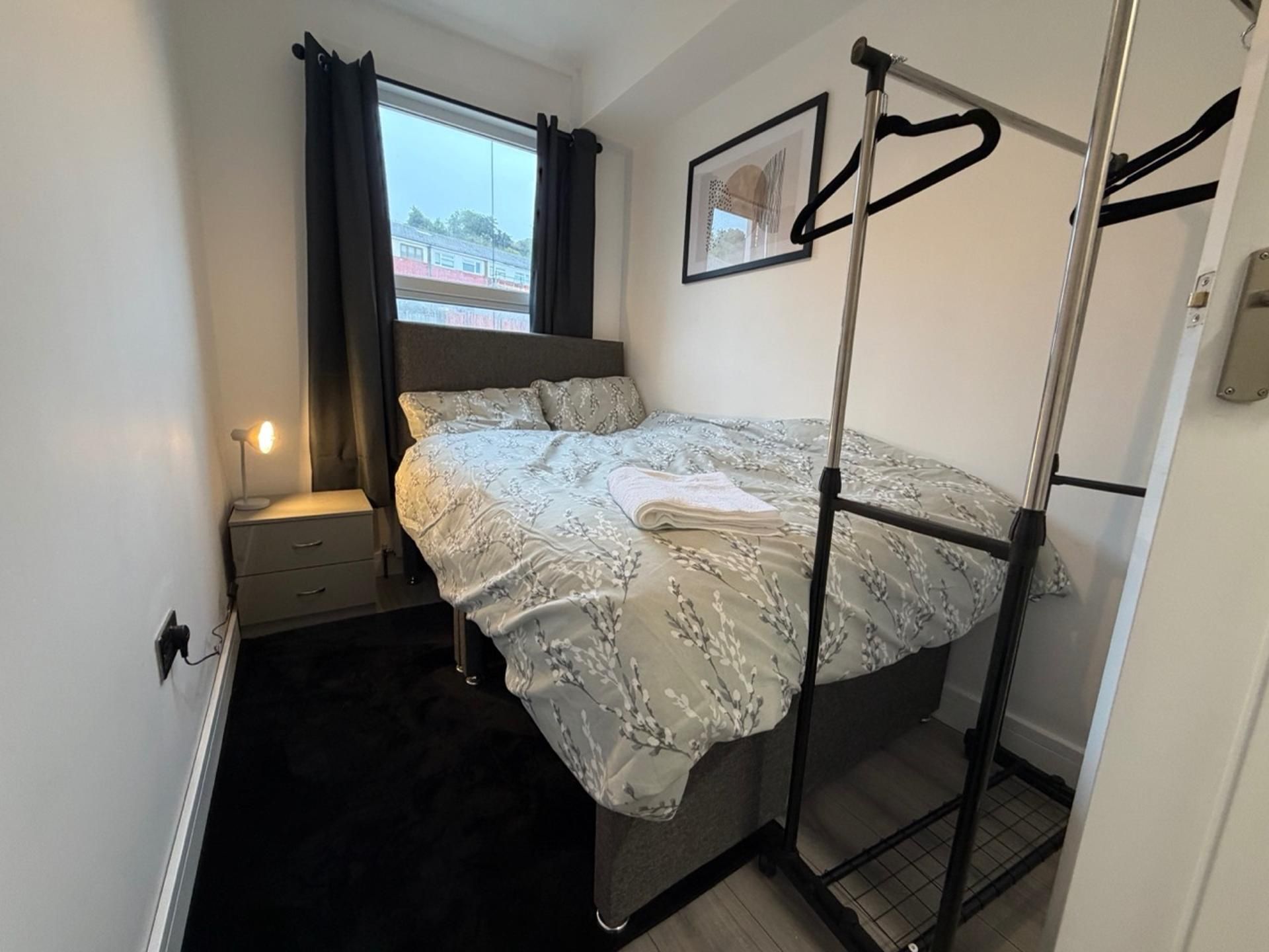 A bedroom with a bed, a nightstand, a window with curtains, and a wall-mounted coat rack.