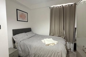 Ein Schlafzimmer mit einem Bett, einem Nachttisch, einem gerahmten Bild an der Wand und einem Fenster mit Vorhängen.