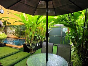 A glass table with chairs under an umbrella, surrounded by lush greenery and a small pool.