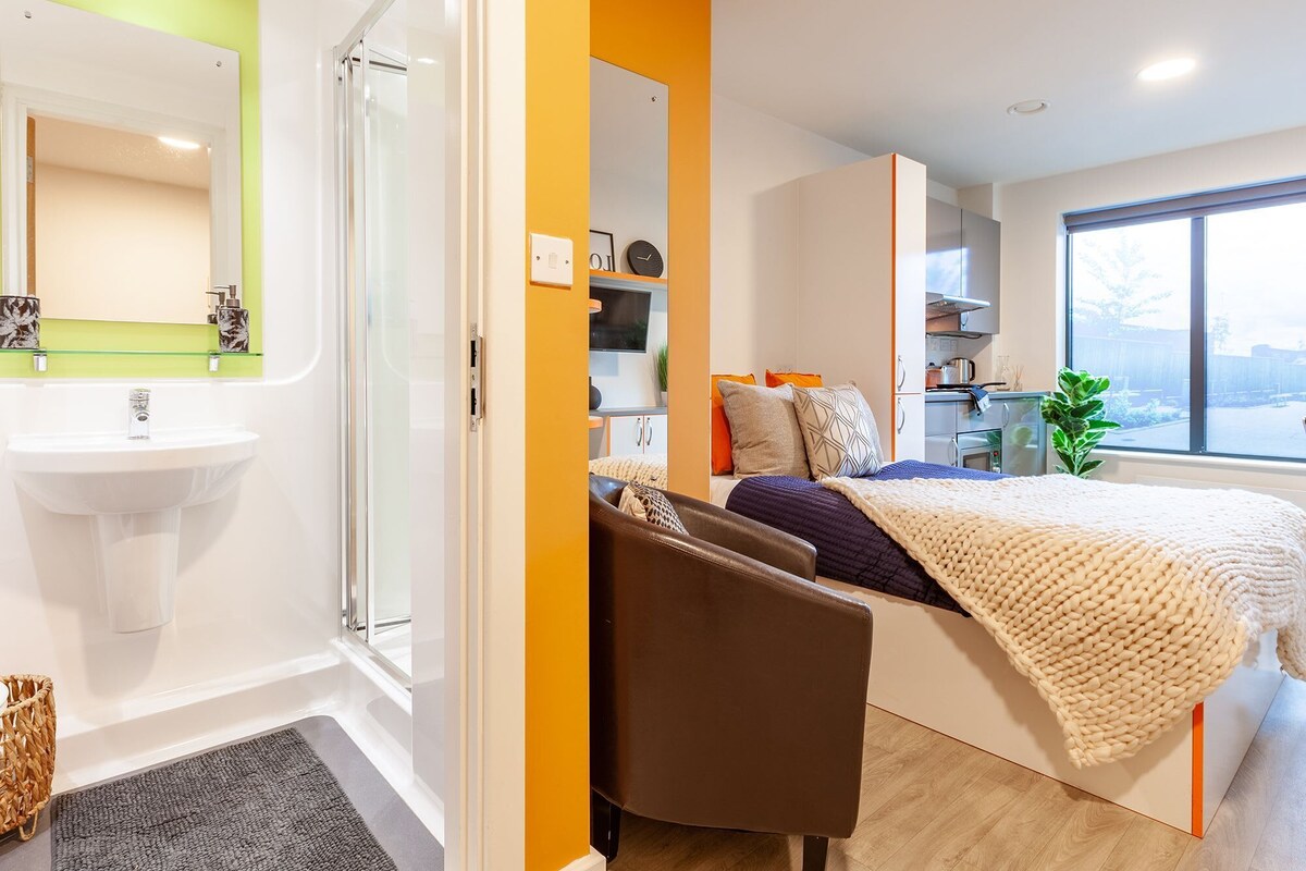 A modern bedroom with a large bed, a bedside table, a chair, a window, and a bathroom with a sink and mirror.